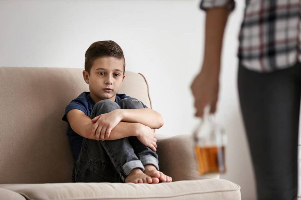 A sad child sitting on a couch looking at his mom holding an alcoholic drink.