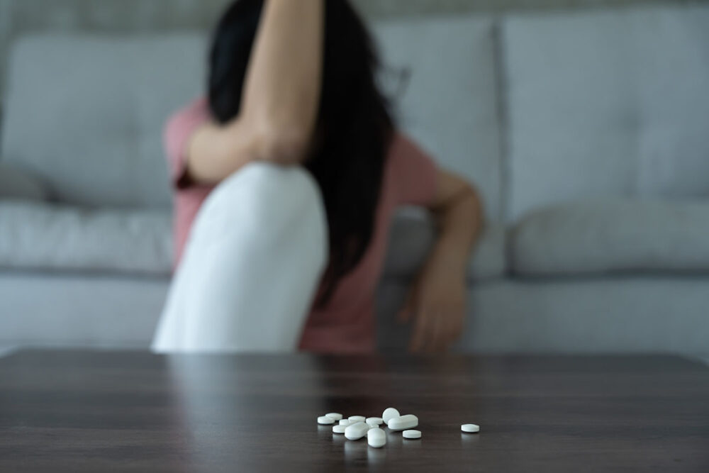 A sad woman with her head down, sitting next to pills on a table.